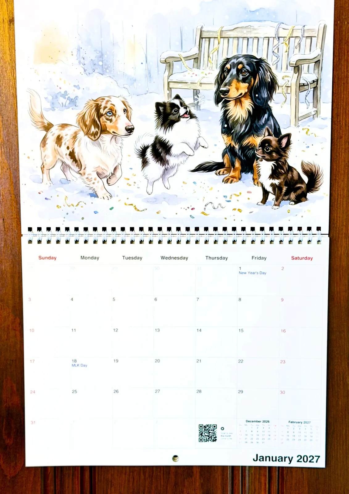 A printed Watercolor calendar hanging on a wooden door, displaying a January page with painted dogs in the snow.