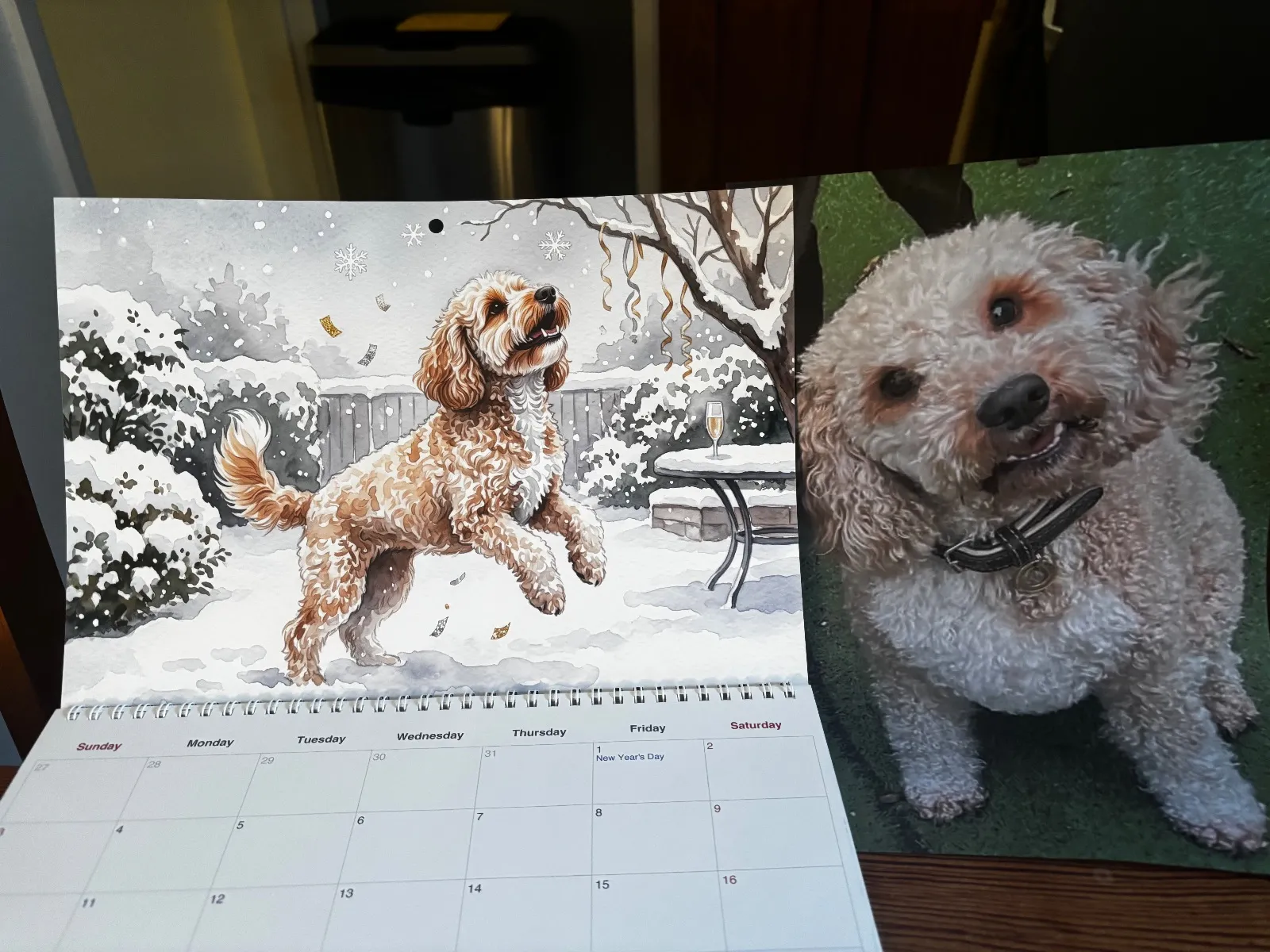 A printed Watercolor calendar opened to a snowy illustration sits next to a photo of the dog.